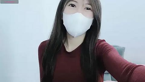 Media: A video of an Asian woman with long black hair, wearing a white surgical mask, red long-sleeve shirt, and seated in a light-colored room.