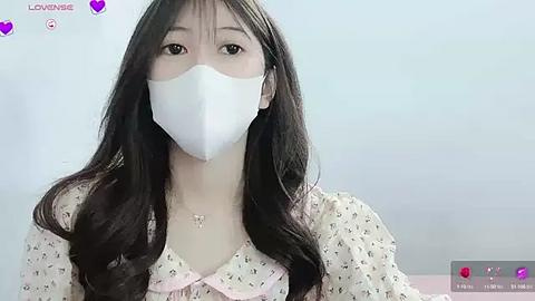 Media: Video of a young Asian woman with long black hair, wearing a white face mask, pink floral dress, and a delicate necklace, against a plain light blue background.