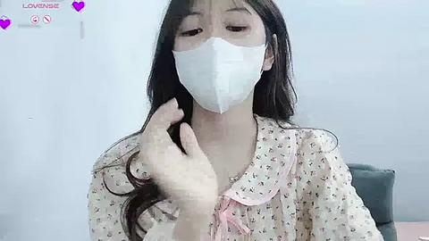 Media: Video of an Asian woman with long black hair in a white floral dress, wearing a white surgical mask, having her hair brushed by an unseen person in a bright, minimalistic room.