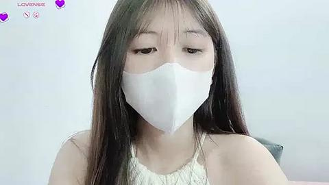 Media: A video of an Asian woman with long, straight black hair wearing a white surgical mask and a white sleeveless top, taken indoors with a plain white background.