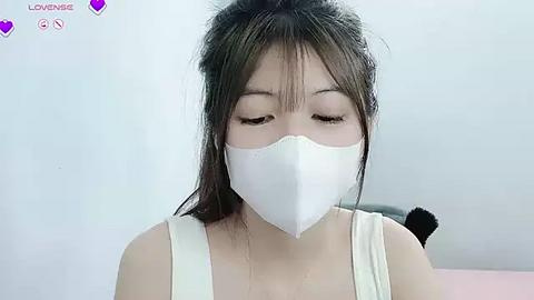 Media: A video of an Asian woman with long, dark hair in a messy bun, wearing a white mask, white tank top, and looking downward.