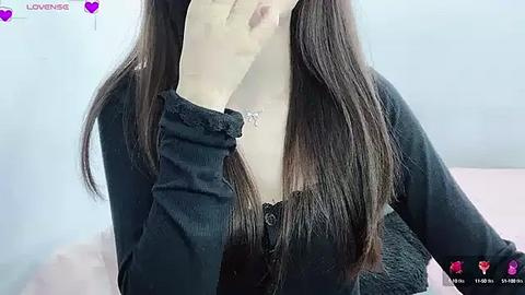 Media: Video of a young woman with long, straight dark hair, wearing a black cardigan, partially covering her face with her right hand. Background shows a blurred white wall and a pink surface.