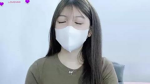 Media: Video of an Asian woman with long black hair, wearing a white surgical mask and olive-green shirt, sitting in a chair against a white background, eyes closed, with \"LOVEVISE\" text at top left.