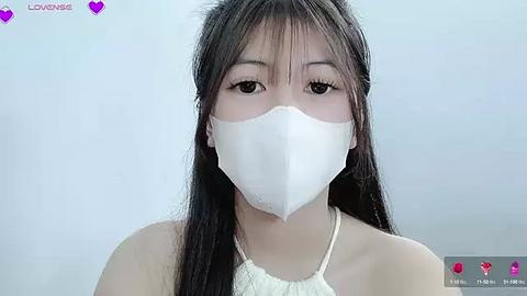 Media: Video of an Asian woman with long black hair, wearing a white surgical mask and a white lace top, against a plain light blue background.