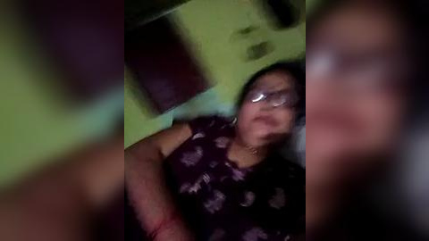 Media: Video of a woman with medium brown skin and short black hair, wearing glasses and a dark floral dress, lying on a bed in a dimly lit room with green walls.