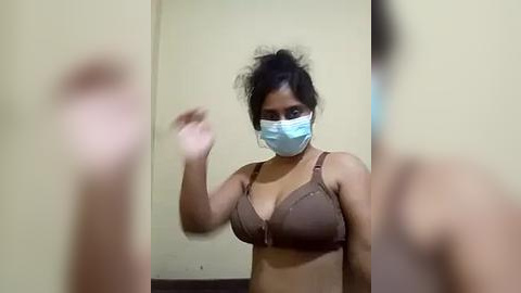 Media: A video of a South Asian woman with medium brown skin, wearing a blue face mask, a brown bra, and a messy bun. She is captured in a dimly lit room with a beige wall and a blurred figure in the foreground.