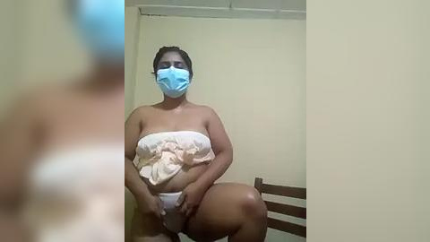 Media: Video of a woman with a medium complexion, wearing a blue face mask and a white cloth covering her breasts, sitting on a wooden chair in a plain room with beige walls.