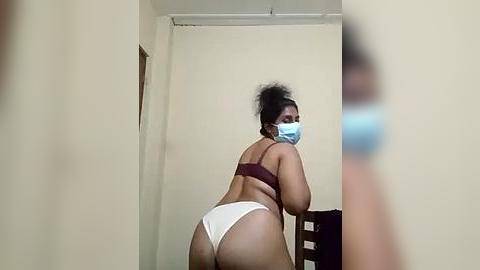 Media: Video of a plus-sized Black woman with dark skin, wearing a maroon bra and white panties, leaning over a chair, face partially obscured by a blue mask, in a plain, beige-walled room.