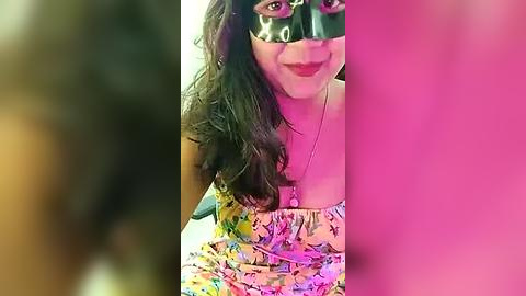 Media: Video of a woman with long black hair, wearing a black Venetian mask and a colorful, floral-patterned skirt, standing in a pink-lit room.