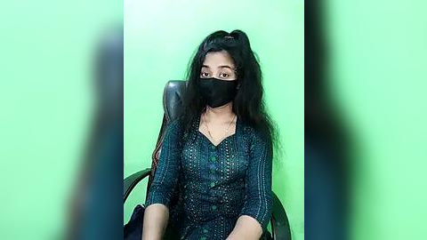 Media: Video of a young woman with long black hair, wearing a green mask, black blouse, and sitting in a black chair against a green screen background.