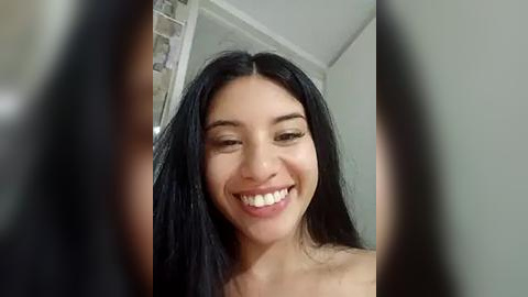 Media: Video of a young woman with long black hair, light skin, and a broad smile. She is indoors, possibly in a bathroom with a shower visible in the background. The image is slightly blurred.