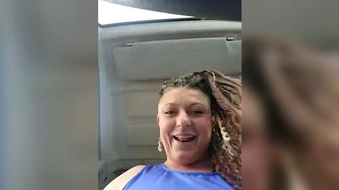 Media: Video of a smiling, light-skinned woman with curly, shoulder-length hair and a blue tank top, sitting in a car with a gray headrest and window in the background.