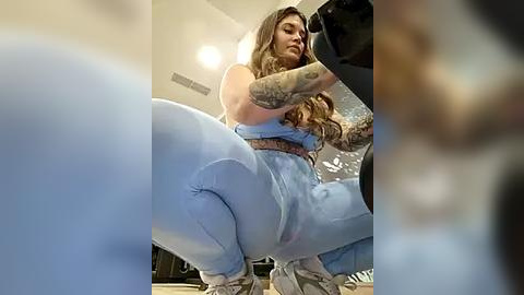 Media: Video of a tattooed woman in a light blue bodysuit, squatting, with a blurry background.