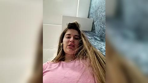 Media: Video of a woman with long brown hair, wearing a pink shirt, standing in front of a waterfall in a modern, light-colored bathroom.