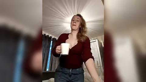 Media: A video of a fair-skinned woman with blonde hair, wearing a red cardigan and blue jeans, holding a white mug in a cozy bedroom with a ceiling fan, gray curtains, and a bed.