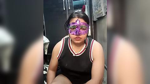 Media: Video of a young woman with medium skin tone and dark hair, wearing a revealing black sports bra, purple mask with a star design, and sitting in a dimly lit, cluttered room with clothing hanging in the background.