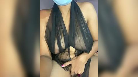 Media: Video of a woman with medium brown skin, wearing a see-through black halter top, exposing her breasts and nipples. She has a blue mask covering her nose and mouth, and her nails are painted red.