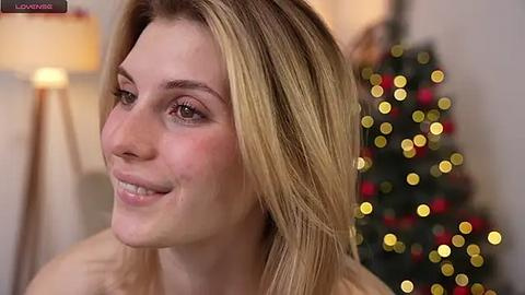 Media: Video of a smiling, fair-skinned woman with long blonde hair, wearing minimal makeup, in a warmly lit room with a Christmas tree in the background.