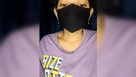 Media: Video of a person wearing a black face mask, a purple shirt with yellow and white text, and dangling earrings, against a dark background.