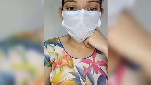 Media: Video of a woman with medium skin tone wearing a white face mask and colorful floral-patterned top. She has dark hair pulled back and her hand rests on her neck. Background is blurred.