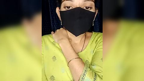 Media: Video of a woman in a yellow saree with black mask, looking down, hands touching her neck, dark background.