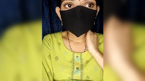 Media: Video of a woman with fair skin, brown eyes, and short black hair, wearing a black face mask, green traditional dress with floral patterns, and a yellow blouse, against a dark blue background.