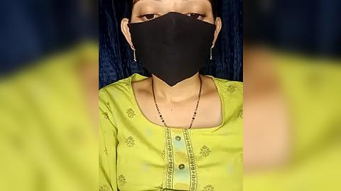 Media: Video of a woman wearing a black face mask, green floral-patterned blouse, and dangling earrings, sitting on a dark blue cushioned chair.