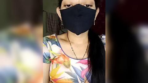 Media: Video of a woman with long black hair, wearing a black face mask and a colorful floral shirt, standing indoors with a blurred background.