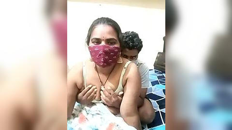 Media: A video shows a woman with a maroon face mask, beige bra, and floral-patterned bedsheet, cradling a baby in a white shirt. Background features a blurred man, bed, and blue-patterned blanket.