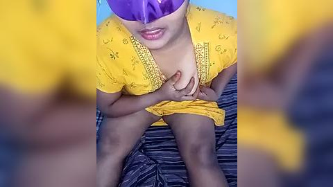 Media: A video showing a partially obscured person with dark skin, wearing a yellow top and purple mask, with hands touching their chest, on a striped blue and black bed.