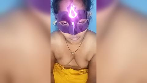 Media: Video of a topless South Asian woman with medium brown skin, wearing a purple mask with a glowing white symbol, and a golden necklace. She's kneeling on a yellow cloth, with a blue background.