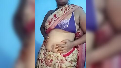 Media: Video of a pregnant South Asian woman in a colorful saree, showcasing a purple bra, hands on her belly, against a blue background.