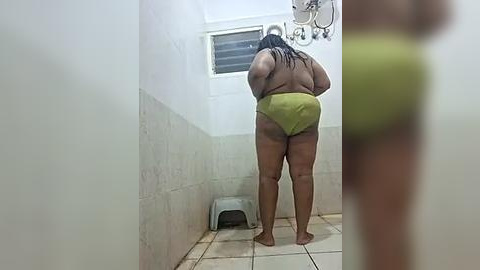 Media: Video of a plus-sized, dark-skinned woman with long black braids in a tiled bathroom, wearing green bikini bottoms, standing barefoot.