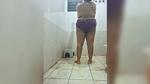 Media: Video of a nude woman with pale skin and short hair, wearing purple bikini bottoms, standing in a tiled shower with white walls and a small step stool.