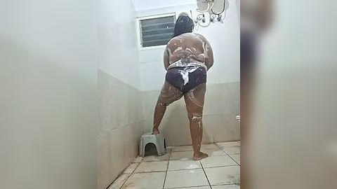 Media: A video shows a dark-skinned woman with a medium build, wearing a dark blue swimsuit, scrubbing a tiled shower stall with a gray plastic brush. The stall is small and clean, with a white window and a metal showerhead.