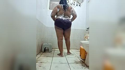 Media: Video of a dark-skinned woman with braided hair in a dingy, tiled bathroom. She's wearing a silver tank top and purple shorts, standing barefoot, facing away from the camera.