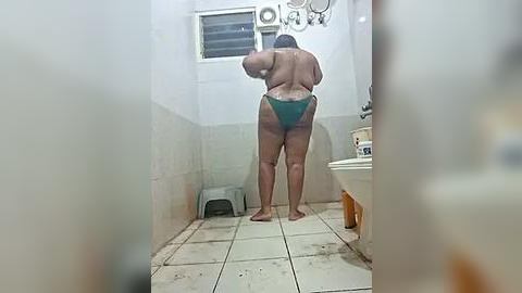 Media: Video of a woman with medium build, wearing green bikini bottoms, standing in a small, cluttered bathroom with tiled walls, white sink, and a toilet.