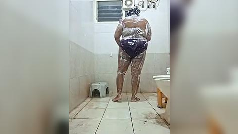 Media: Video of a bare-chested man in a tiled bathroom, covered in white powder, standing near a white sink and a gray plastic stool.