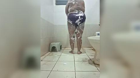 Media: Video of a person, likely a man, showering in a small, tiled bathroom. The subject is facing away from the camera, wearing only dark blue boxer shorts. A green plastic stool is in the corner.