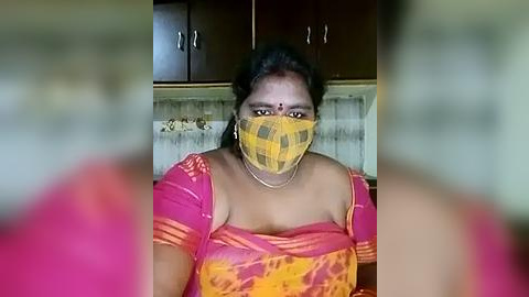 Media: Video of a South Asian woman in a bright yellow and orange saree, wearing a checkered face mask and a bindi, standing in a kitchen with dark wooden cabinets and a tiled backsplash.