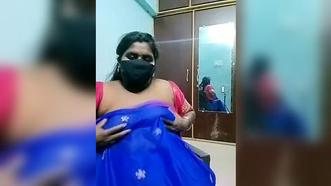 Media: A video of a woman in a blue saree with a black face mask, sitting in a brightly lit room with wooden cupboards and a mirror, focusing on her reflection.