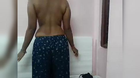 Media: Video of a topless person with medium brown skin, wearing navy blue pajama pants with small white patterns, standing in a dimly lit room with light pink walls and a white tiled floor.