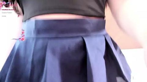 Media: Video of a woman wearing a short-sleeved, black crop top and a pleated, navy blue skirt with a small, decorative bow. The background is blurred, emphasizing her attire.