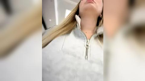 Media: A close-up video of a blonde woman in a light grey, zip-up hoodie, with her head tilted back, showing her neck and part of her face. The background is blurred, suggesting an indoor setting.