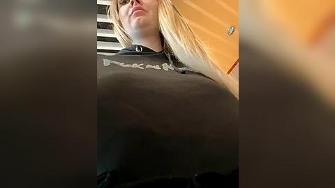 Media: Video of a blonde woman with long hair, wearing a black hoodie, taken from a low angle, indoors, with blurred background.