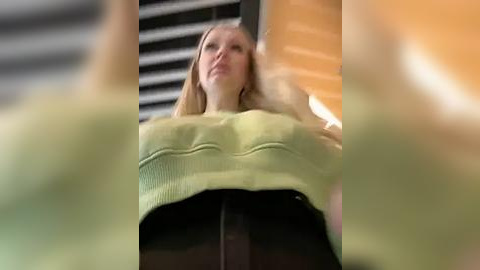 Media: A blurred, low-angle video shows a blonde woman in a green sweater and black pants, her face partially visible, with a wooden ceiling and horizontal blinds in the background.