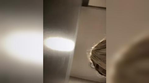 Media: Video of a blonde woman with her hair tied back, illuminated by a bright, circular light source, creating a dramatic contrast against a dark, metallic surface.