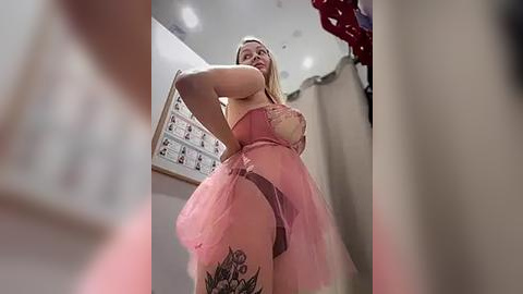 Media: Video of a curvy, blonde woman in pink lingerie with a floral tattoo on her thigh, standing in a dimly lit room with a whiteboard and curtains in the background.