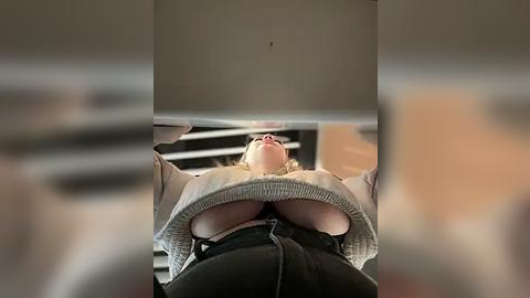 Media: Video of a woman in a gray sweater and black pants, lying on her back, looking up at the ceiling, with a blurred background featuring furniture and light-colored walls.