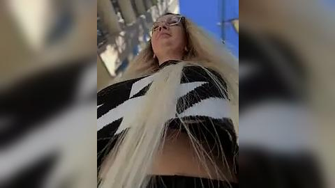 Media: A video of a woman with long blonde hair, wearing glasses and a black and white striped top, standing in a blurred, urban setting with blue sky and buildings.